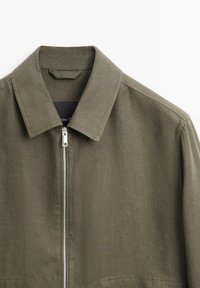 Olivengrøn jakke med spids krave og frontlynlås i sølv, vist på hvid baggrund.