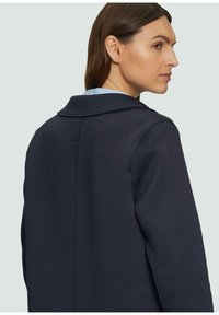 Cappotto blu navy con un ampio colletto, linee pulite e dettaglio cucitura sul retro discreto, realizzato in un tessuto liscio con una vestibilità personalizzata.