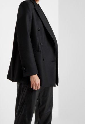 Personne portant un blazer noir ample à double boutonnage et un pantalon en cuir noir, visible de l'épaule jusqu'à mi-cuisse sur un fond uni.