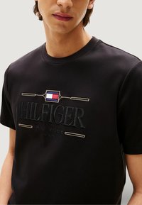 Maglietta nera a maniche corte con la scritta "HILFIGER NEW YORK 1985" ricamata in filo scuro, con un piccolo dettaglio logo rosso, bianco e blu.