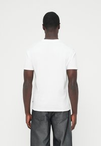 Maglietta bianca in cotone con maniche corte, collo rotondo e trama liscia, vista da dietro su sfondo chiaro.