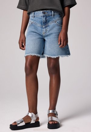 Shorts di jeans azzurri con orlo sfrangiato, dotati di chiusura con bottone e cerniera. Sandali argentati con suola nera piatta e cinturini metallici.