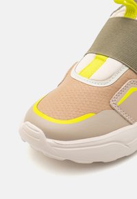 Αθλητικά παπούτσια με υφή από μπεζ διχτυωτό υλικό, παχύ λευκό σόλα και νέον κίτρινες λεπτομέρειες στην μύτη και στο λουρί. Πράσινη ελαστική ταινία που περιτυλίγει.