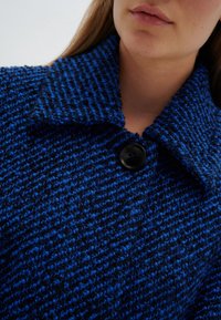 Kvinna som bär en strukturerad blå och svart vävd kappa med en stor krage och en enda svart knapp f äst nära halsen.