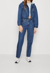 Džínová bunda s kapucí, světle modrý krátký top a modré džíny s rovnými nohavicemi. Bílé tenisky se šedými,  černými a oranžovými detaily.