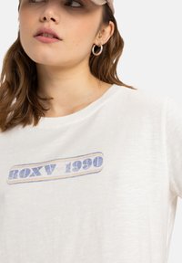 Balta trumpo rankovėmis marškinėliai su daugiakopiu „ROXY 1990“ piešiniu, malonios tekstūros ir laisvo kirpimo.