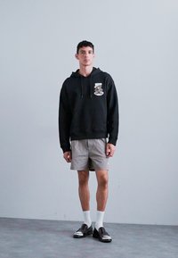 Schwarzer Hoodie mit Frontgrafik, graue karierten Shorts, weiße Knöchelsocken und schwarze Schuhe mit glänzendem Finish. Person steht vor einem hellen Hintergrund.