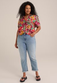Kurzärmliges Hemd mit einem lebhaften Blumendesign in Rot, Gelb und Grün auf einem weinroten Hintergrund; kombiniert mit hellblauen, umgeschlagenen Jeans und schwarzen Sandalen.