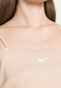 Hladký béžový top na ramínka s jemným žebrováním, který má malé bílé logo Nike uprostřed přední části. Hladká textura s minimálními detaily.