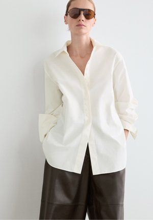 Femme portant une chemise crème oversize boutonnée, un pantalon en cuir large et foncé, et des lunettes de soleil marron devant un mur blanc uni.