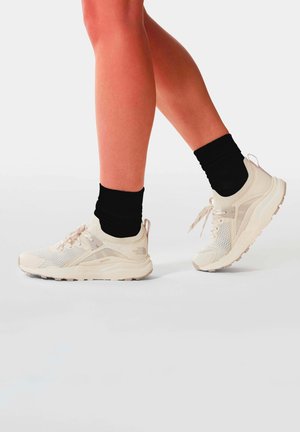 Jambes portant des chaussettes noires courtes et des baskets en mesh beige The North Face sur fond blanc, un pied légèrement levé.