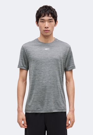 Junger Mann trägt ein graues Sport-T-Shirt mit kurzen Ärmeln und einem kleinen weißen Logo auf der Brust, steht vor einem schlichten Hintergrund.