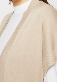 Cardigan di maglia beige con bordo a coste, design aperto sul davanti e una texture leggera. Indossato sopra una maglietta bianca. Design semplice e contemporaneo.