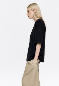 Camiseta negra de gran tamaño con mangas cortas, adornada con tachuelas metálicas esparcidas por toda la prenda, combinada con pantalones sueltos de color beige.