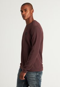 Pulover tricotat cu mâneci lungi de culoare burgundy, cu un finisaj texturat, decolteu rotund și un mic logo negru pe piept, asortat cu blugi de culoare albastră deschisă.