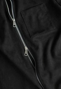 Sorte semsket stoff har en sølvglinsende glidelås langs kanten, med en kvadratisk lomme synlig på overflaten, noe som skaper et stilrent, teksturert design.