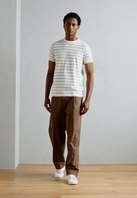 White striped t-shirt with short sleeves, paired with brown loose-fit trousers and white sneakers, standing on a wooden floor.