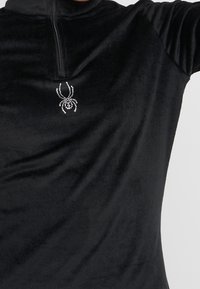 Pulóver de forro polar negro con cuello con cremallera, que presenta un diseño de araña brillante en rhinestones en el pecho. Textura suave.