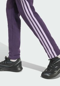 Fialové tepláky s bielymi bočnými pruhmi, kombinované s čiernymi športovými topánkami s čiernymi a šedými akcentmi a sieťovinovými detailmi.