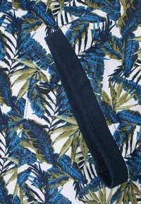 Tropisk-mønstrede stof med grønne og blå blade på en hvid baggrund; et mørkt elastisk bånd krydser designet.