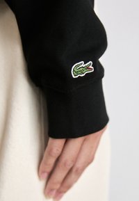 Chemise noire à manches longues avec un logo de crocodile vert sur le poignet, fabriquée en tissu texturé, avec un design ajusté.