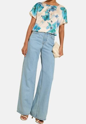 Femme portant un chemisier floral crème, un jean large bleu clair, des sandales blanches à talons, et tenant une petite pochette crème.