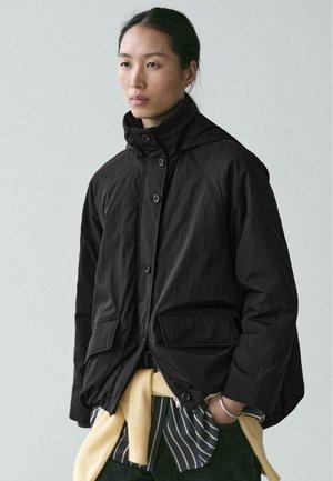 Veste noire à col montant, fermeture à boutons, deux poches avant, et coupe décontractée. Portée sur une chemise rayée et un pull noué.