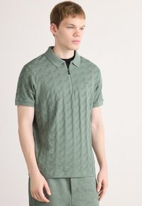 Ung man klädd i en kortärmad polo t-shirt med grönt geometriskt mönster och en dragkedja framtill, samt matchande shorts, som tittar åt sidan.