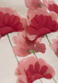 Textiel met gelaagde rode en roze stoffen bloemen op een crèmekleurige achtergrond, met groene stelen en fijne stikaccenten.
