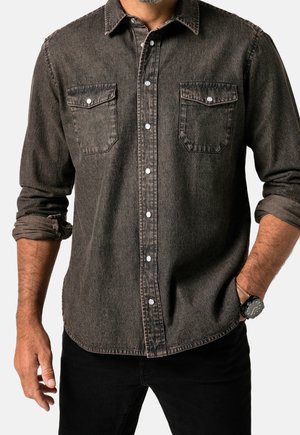 Mann trägt ein verblasstes schwarzes Denim-Hemd mit Knöpfen und hochgekrempelten Ärmeln, schwarze Hose und eine schwarze Armbanduhr am linken Handgelenk.