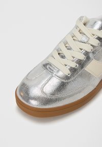 Sneaker con finitura crackle argentata, con lacci crema e un accento laterale beige, caratterizzato da una punta tonda e una suola in gomma color caramello.