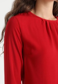Top rosso a maniche lunghe con scollatura rotonda e un sottile dettaglio a plissettature. Realizzato in tessuto liscio, con una silhouette aderente.