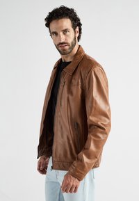 Man met krullend haar en baard die een bruine leren jas draagt over een zwart shirt en lichte spijkerbroek, staand tegen een effen achtergrond.