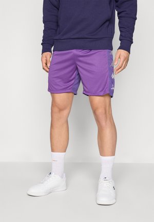 Uomo che indossa pantaloncini sportivi viola, calzini bianchi, scarpe da ginnastica bianche e una felpa blu scuro in piedi su un pavimento grigio davanti a una parete bianca.