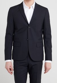 Navy-blaues, maßgeschneidertes Sakkos in einem glatten Stoff, mit einem Zwei-Knopf-Verschluss, Reverskragen und zwei vorderen Taschen. Getragen über einem weißen Hemd.