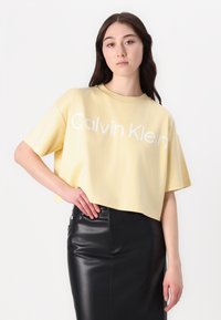 Gul croppet t-skjorte med hvit "Calvin Klein" logo, korte ermer, rund hals; kombinert med et svart skinn blyantskjørt.