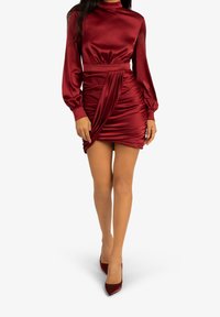 Czerwona satynowa sukienka z długim rękawem i wysokim dekoltem, z marszczeniami w talii i drapowaną spódnicą; zestawiona z dopasowanymi czółenkami na obcasie.