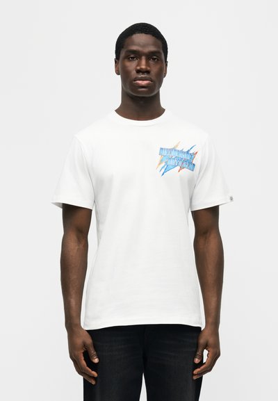 Άνδρας μοντέλο που φοράει ένα λευκό T-shirt με πολύχρωμο λογότυπο "Billionaire Boys Club" στην αριστερή πλευρά του στήθους, συνδυασμένο με σκουρόχρωμο παντελόνι, στέκεται σε απλό φόντο.