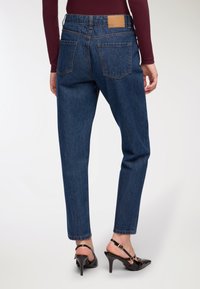 Osoba ubrana w ciemnoniebieskie spodnie z wysokim stanem, burgundową bluzkę z długim rękawem i czarne sandały na obcasie, pokazana od tyłu.