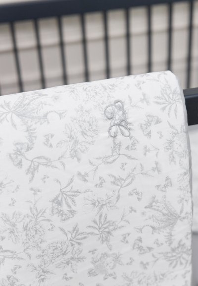 Λευκό ύφασμα με περίτεχνο γκρι ανθικό σχέδιο, που διαθέτει κεντημένη διακοσμητική λεπτομέρεια. Απαλή υφή, απλωμένο πάνω σε κάγκελο κούνιας.