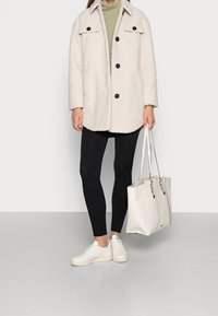 White fuzzy jacket with buttons, green top, black leggings, and white sneakers. A beige handbag with silver hardware is held in one hand.