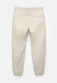Beige byxor med resårmidja, avsmalnande ben och en bakficka med en vikt detalj. Slät, mjuk textur.