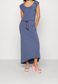 Niebieska sukienka maxi z krótkimi rękawami, dekoltem w łódkę oraz splecionym paskiem w talii. Posiada kieszeń i jest zestawiona z różowymi sandałami na platformie w kratkę.