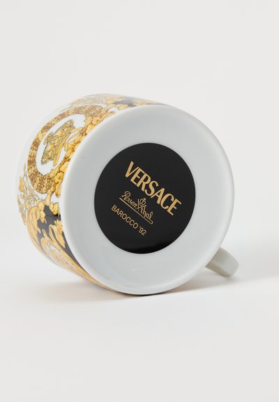 Κεραμική κούπα με γυαλιστερή λευκή βάση, περίπλοκο χρυσό σχέδιο Barocco και μαύρο κυκλικό αυτοκόλλητο που υποδεικνύει την επωνυμία και το μοντέλο.