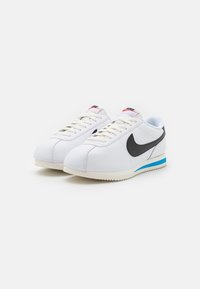 Sneakers in pelle bianca con uno swoosh nero, un accento blu sul tallone e una suola in gomma testurizzata. Presentano lacci bianchi e branding sulla linguetta.