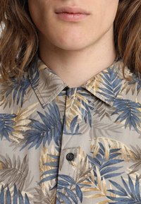 Short-sleeve shirt in a light beige fabric with blue and gold leaf pattern, featuring a button-down collar and a single button closure at the neck.