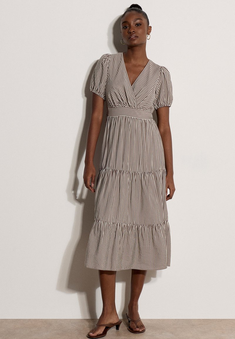 Femme portant une robe midi à rayures verticales marron et blanche avec des manches bouffantes et un col en V, associée à des sandales à talons marron.