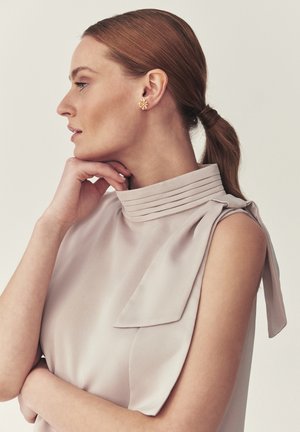 Frau mit zurückgebundenen roten Haaren, trägt ein ärmelloses beiges Oberteil mit plissiertem Kragen und goldenen floralen Steckerohrringen, ruht mit dem Kinn auf der Hand im Profil.