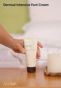 Tuba Dermud Intensive Foot Cream, biała z ciemną podstawą, zawiera 100 ml. Ręka trzymająca tubę obok świecy na drewnianej powierzchni.