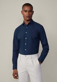 Camisa de manga larga azul marina con cuello puntiagudo, que presenta botones de color claro y un logo sutil. Combinada con pantalones de color claro.
